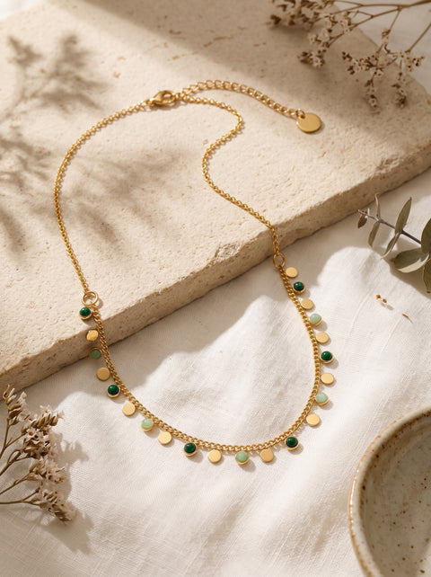A delicate gold chain necklace with small gold disc charms and alternating green and pale green bead accents, styled on a textured beige stone slab with dried botanicals.