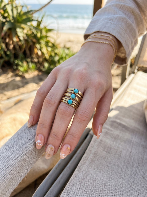 A close-up of a hand wearing a stack of three gold rings, each set with a small round turquoise stone, resting on a beige cushion with a beach and ocean blurred in the background.