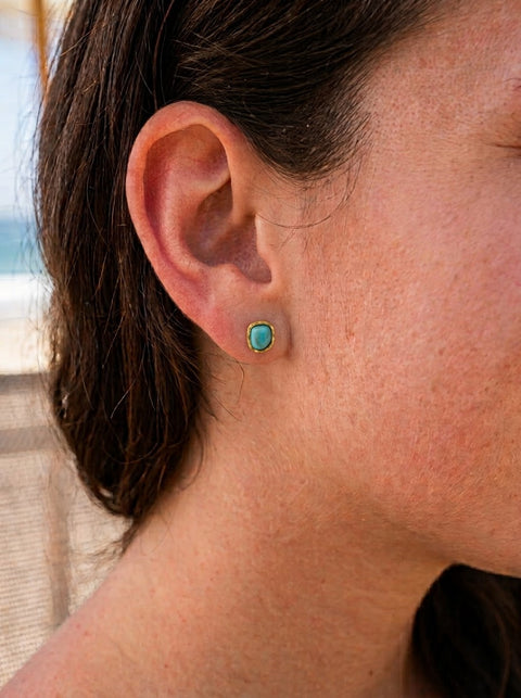 A close-up of a person's ear wearing a small stud earring with a round turquoise-like blue stone in a gold-toned setting.