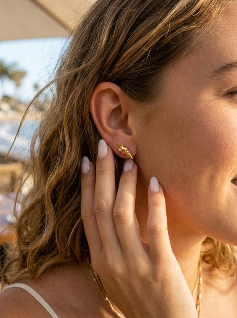 A close-up lifestyle shot of a woman wearing a small, textured gold auger shell stud earring. The photo is taken in warm, sunny outdoor lighting with a blurred beach or resort background.