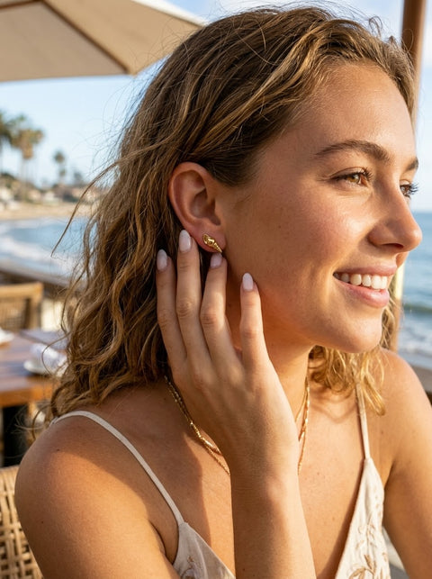 A woman with wavy light brown hair is shown in a close-up profile, wearing a small gold auger shell stud earring and a delicate gold chain necklace in a sunny outdoor setting near the beach.