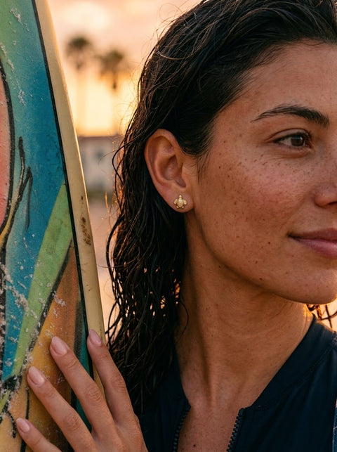 A close-up lifestyle shot of a woman with wet dark hair wearing a small gold sea turtle studs, leaning against a colorful surfboard at sunset.