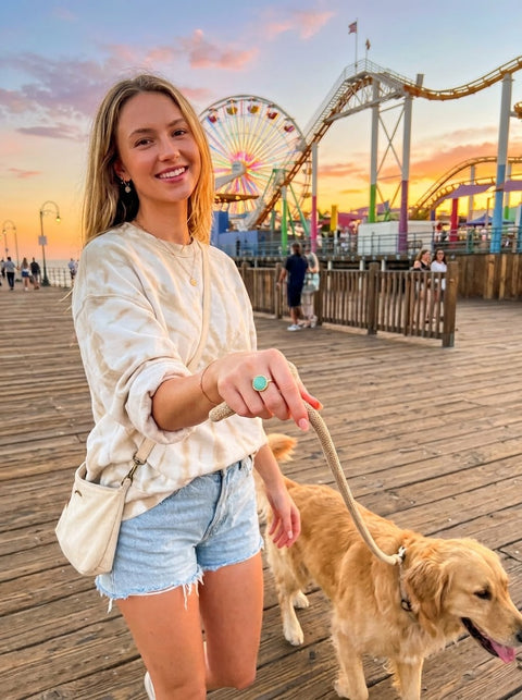 A woman smiling on a wooden boardwalk at sunset while holding a golden retriever on a leash. She is wearing a cream tie-dye long-sleeve top, denim shorts, and a small cream shoulder bag and a Amazonite ring, with a pier and roller coasters in the background.