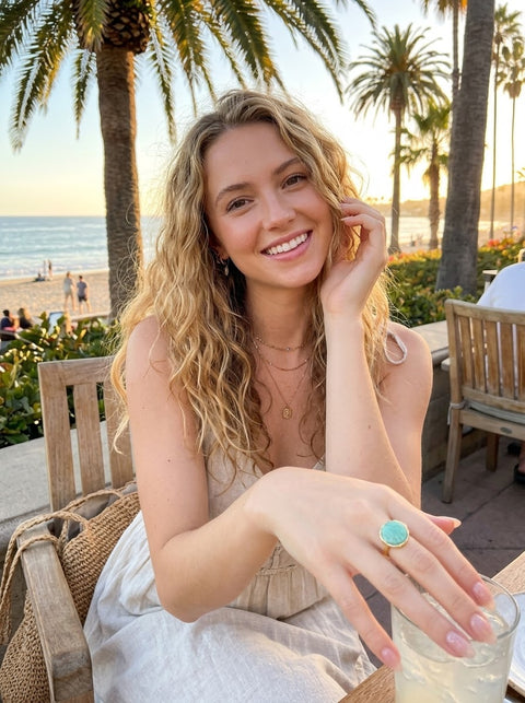 A smiling woman with blonde curly hair sits at an outdoor beachside cafe, wearing a white sleeveless dress and a Amazonite ring on her finger. Palm trees and the ocean are visible in the background.