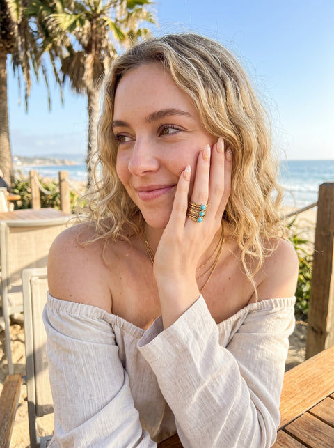 A woman with blonde curly hair wearing a beige off-the-shoulder top and a gold ring with turquoise-colored stones, posing outdoors with a beach and palm trees in the background.