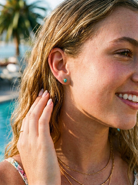 A close-up lifestyle shot of a woman with blonde hair wearing a small turquoise stud earring and layered gold necklaces, set against a sunny outdoor background with a swimming pool and palm trees.
