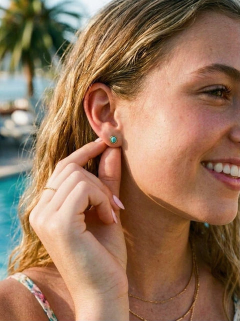 A close-up lifestyle shot of a woman with blonde hair wearing a small turquoise stud earring by a poolside background.