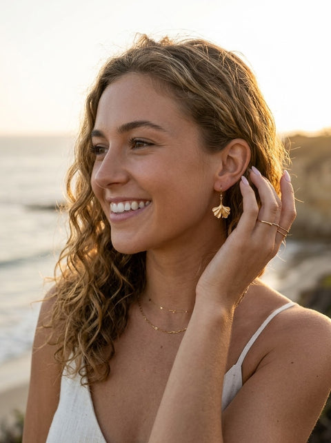 A woman with wavy light brown hair smiling outdoors near the ocean at sunset, wearing a gold gingko drop earring and delicate gold necklaces.