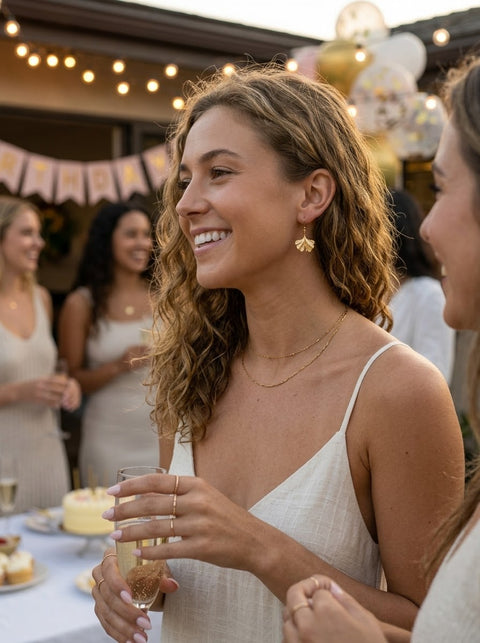 A woman at a party wearing a white spaghetti-strap dress with golden gingko leaf drop earrings, holding a glass of champagne, with other guests and festive decorations in the background.