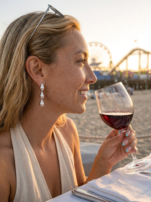 A woman in a beach setting at sunset holds a glass of red wine, wearing silver teardrop earrings and a cream halter top. A Santa Monica Pier scene is visible in the background.