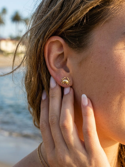 A close-up of a woman's ear wearing a small gold crab stud earring, with her hand resting near her face against a blurred beach background.
