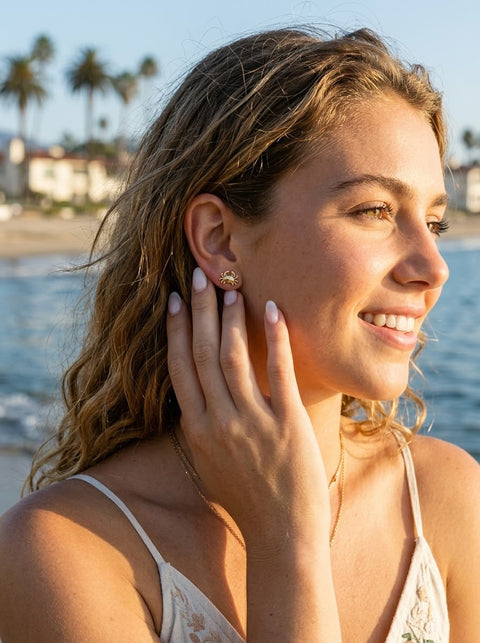 A woman with blonde hair stands on a beach, touching her ear where a small gold crab stud earring is worn. Palm trees and the ocean are visible in the background.
