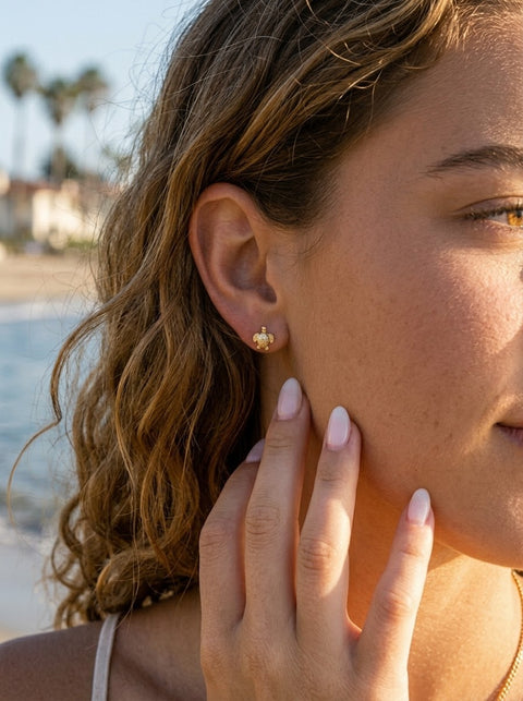 A close-up lifestyle shot of a woman with wavy light brown hair wearing a small gold sea turtle stud earring, set against a blurred beach background.