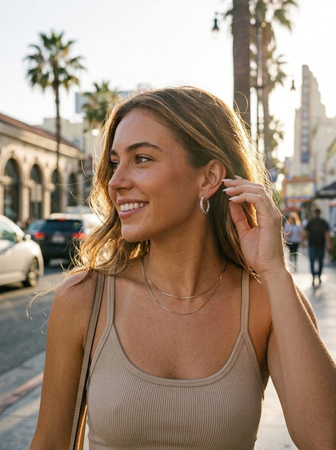 A woman wearing a beige ribbed tank top and layered gold necklaces and a silver twist hoops, smiling on a sunny city street with palm trees in the background.