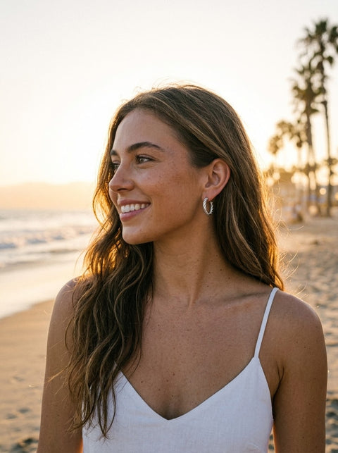 A woman with long brown hair smiling on a beach, wearing a white spaghetti-strap top and small silver hoop earrings, with ocean waves and palm trees in the background.
