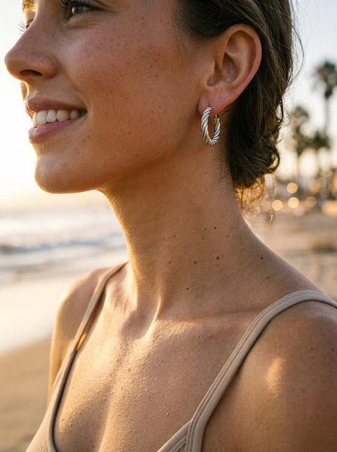 A woman wearing a silver twisted hoop earring, photographed in profile on a beach at sunset. The background shows a blurred shoreline with palm trees and warm golden light.