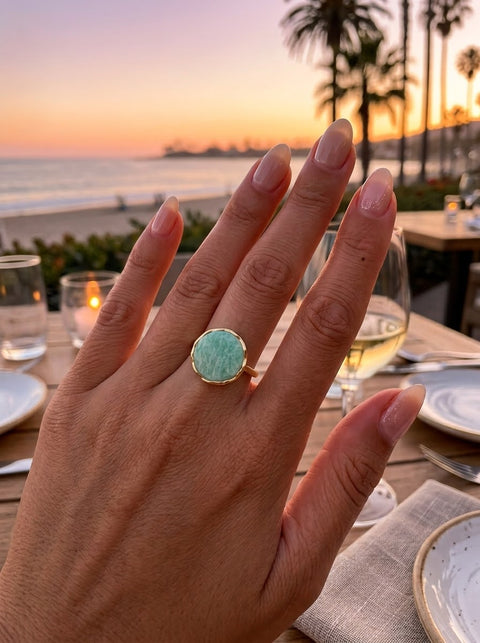 A gold ring with a round light green Amazonite stone worn on a hand, photographed at a beachside restaurant during sunset with plates, wine glass, and palm trees in the background.