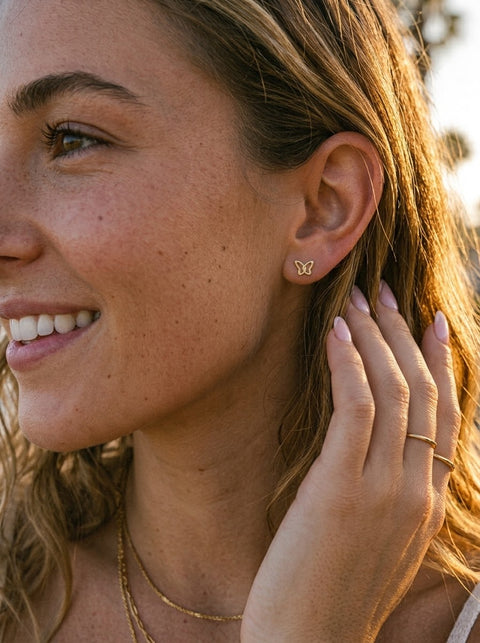 A close-up lifestyle shot of a woman with warm golden-hour sunlight, wearing a small gold butterfly stud earring and delicate gold necklaces.