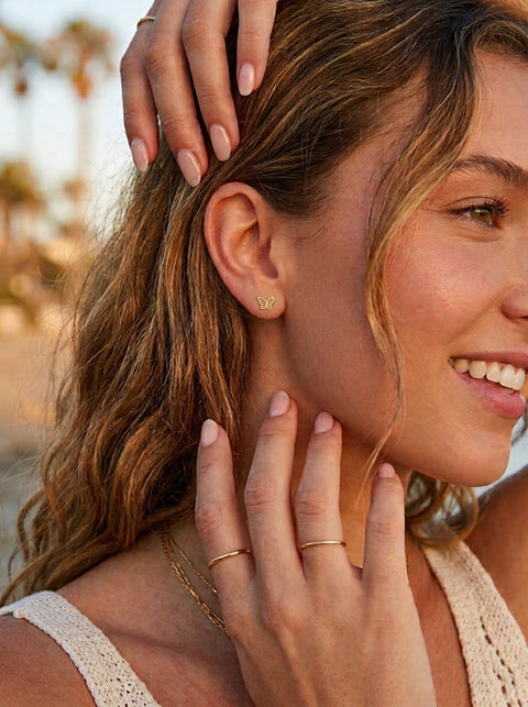 A close-up lifestyle shot of a woman with wavy light brown hair, wearing a delicate gold butterfly-shaped stud earring, a thin gold ring on her finger, and a gold chain necklace. She is smiling and posing outdoors with a warm, blurred background.
