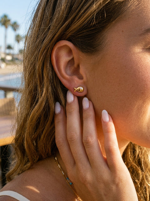 A close-up lifestyle shot of a woman wearing a small gold fish-shaped stud earring, with her hand resting near her ear. The background shows a softly blurred outdoor beach setting with palm trees and water.